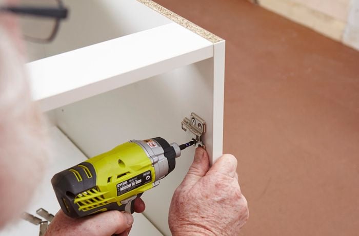 Person drilling screw into kitchen cabinet door hinge.