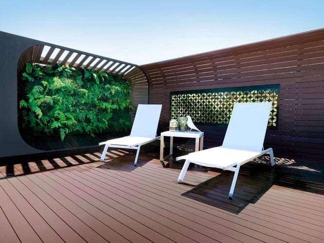 Modern outdoor patio with two white lounge chairs and a small table, set against a vertical garden with lush greenery and wooden panel walls.