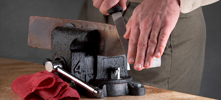 Close-up of a person using a hand file to smooth the edge of a metal piece secured in a bench vice on a wooden workbench, with a red cloth nearby.