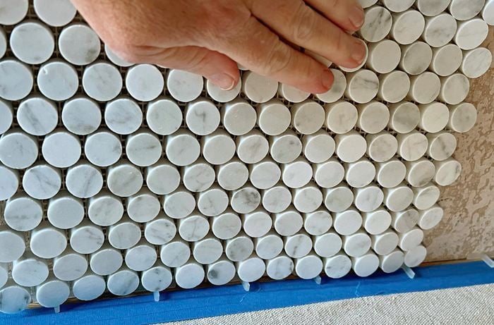 A person positioning a round mosaic tile sheet on a splackback.
