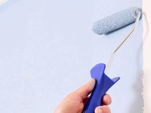 A hand holding a blue Monarch mini paint roller, painting pale blue paint onto a white wall