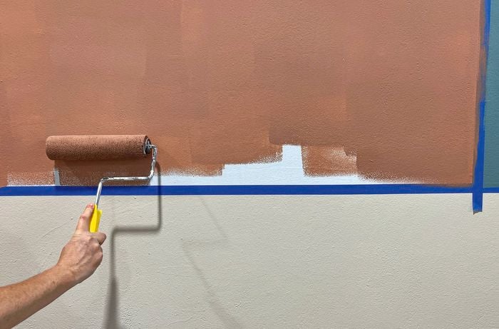 A hand holding a paint roller rolls Taubman’s Baby Bear coloured paint on a white wall, with blue painter’s tape used for colour blocking.
