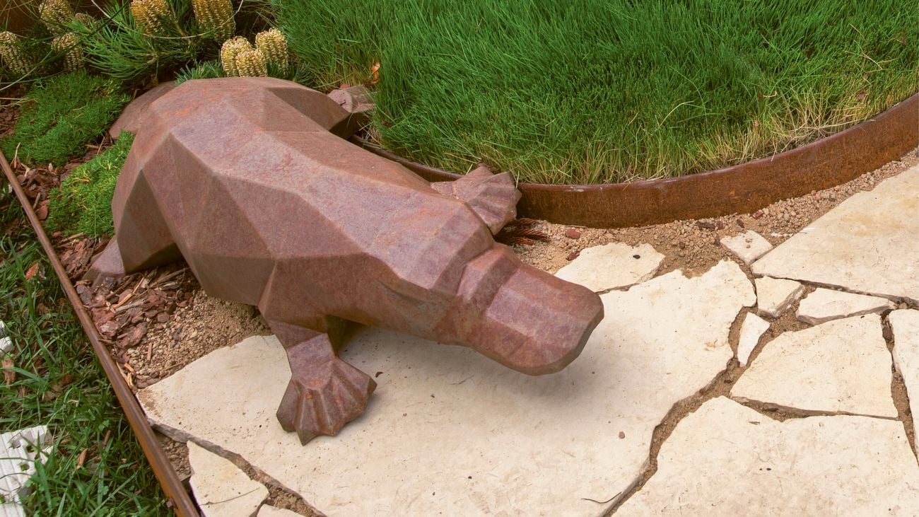 Rust-coloured metal platypus sculpture in a garden bed.