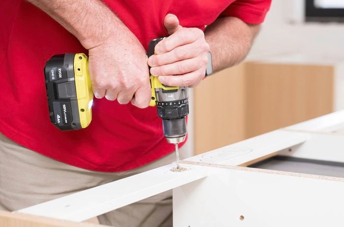 A person is using a power drill to drill a hole into the support rail of the cabinet. 