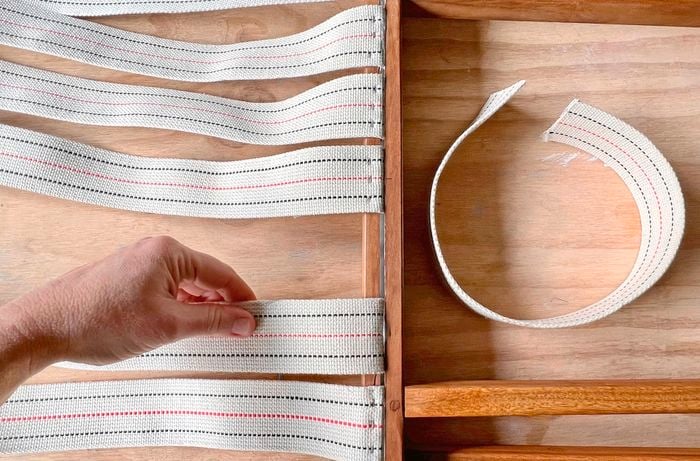 A hand attaching strips of webbing into the groove of a wooden frame with adhesive.
