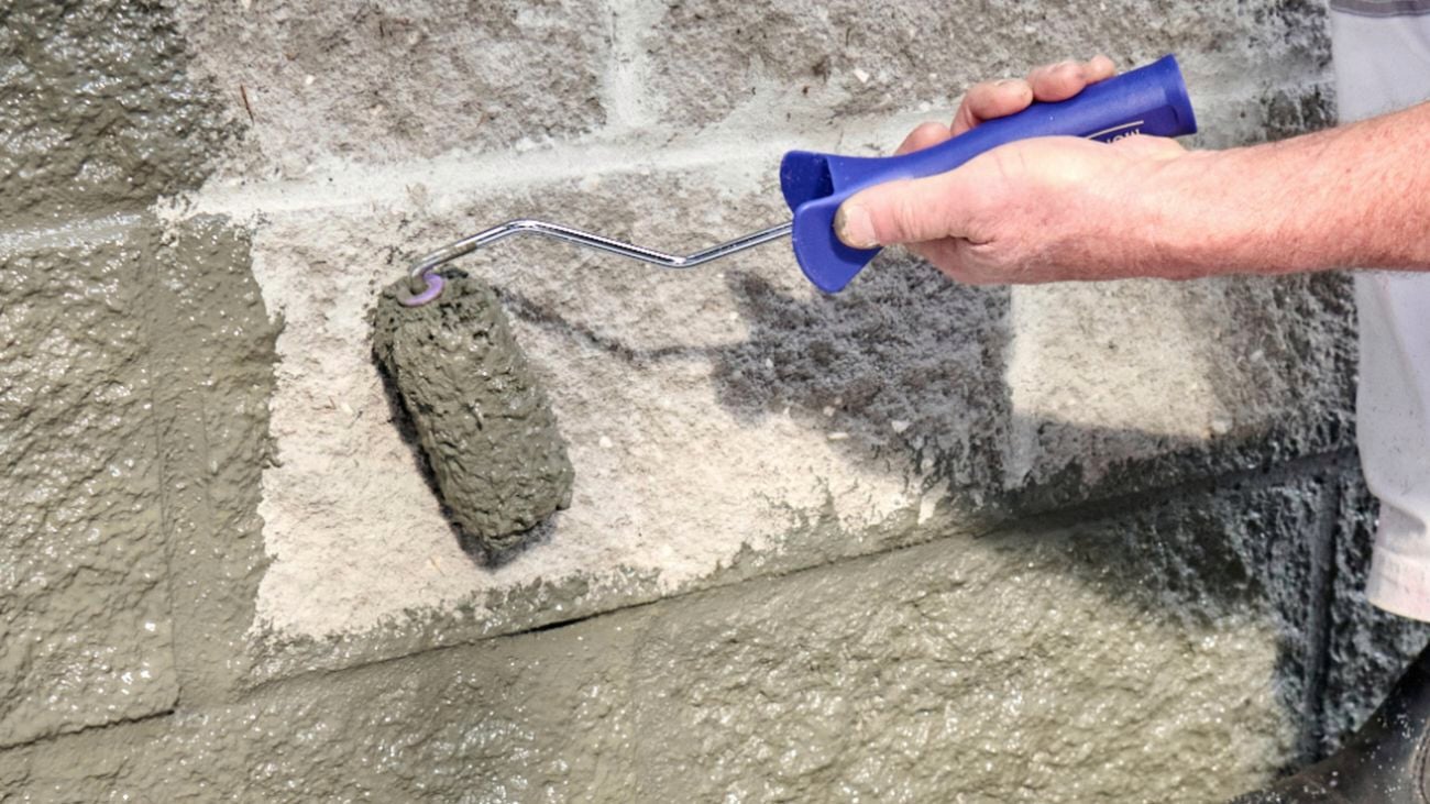 A hand holding a blue Monarch mini paint roller, painting grey paint onto a concrete block wall