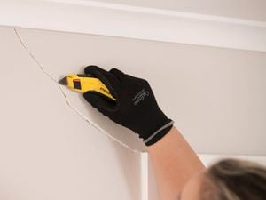 A hand wearing a black safety glove uses a utility knife to repair a cracked wall surface.