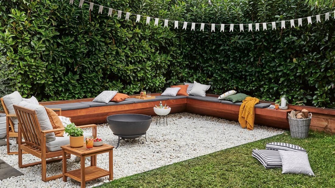 Timber lounge setting with cushions on it, chairs and a fire pit outside on pebbles.