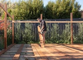 A carpenter in a black t-shirt and light-coloured shorts stands on a half-finished outdoor deck examining decking material - timber.