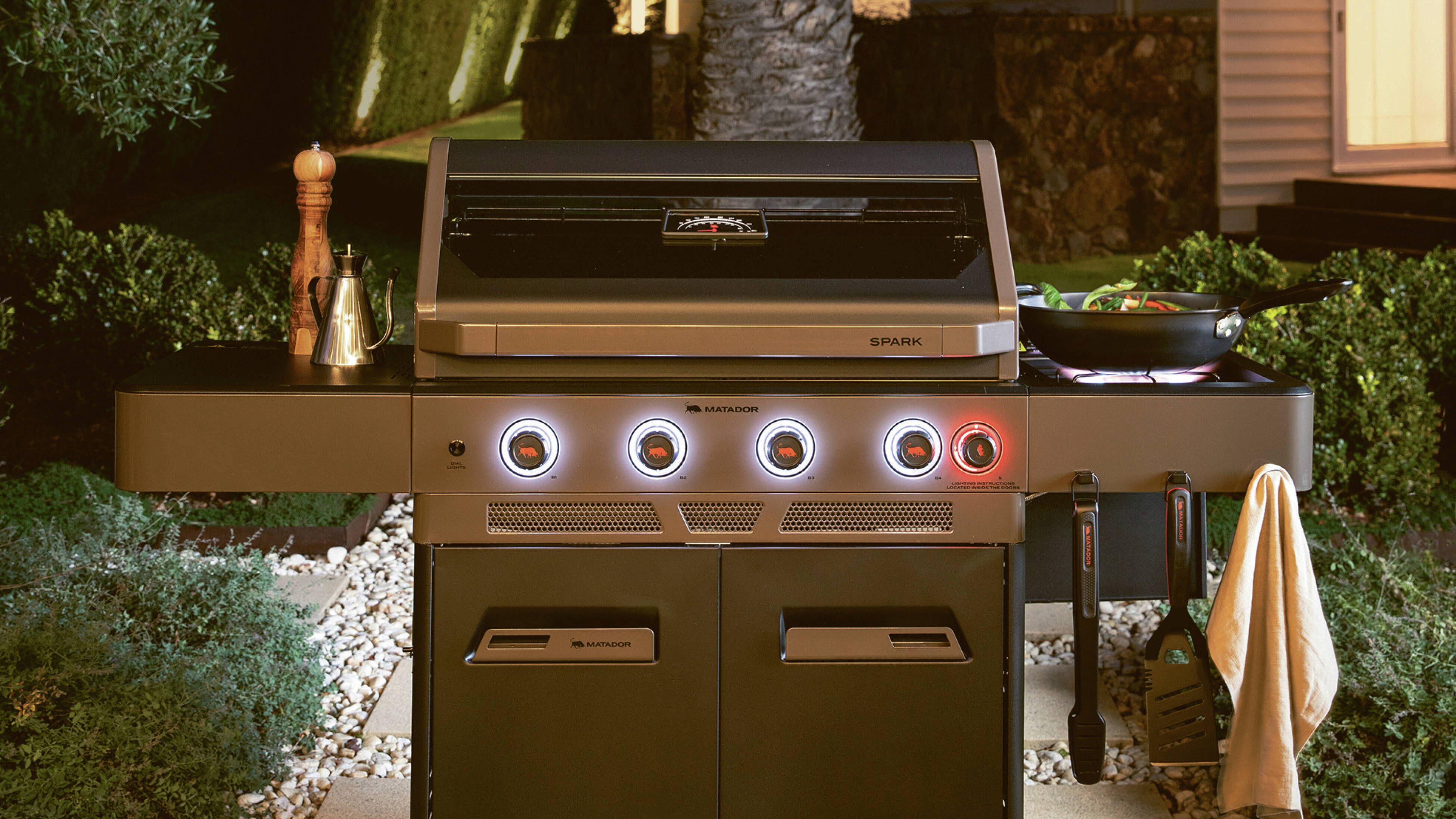 Outdoor barbecue with a pan on the side burner, on a patio with a tree and greenery nearby