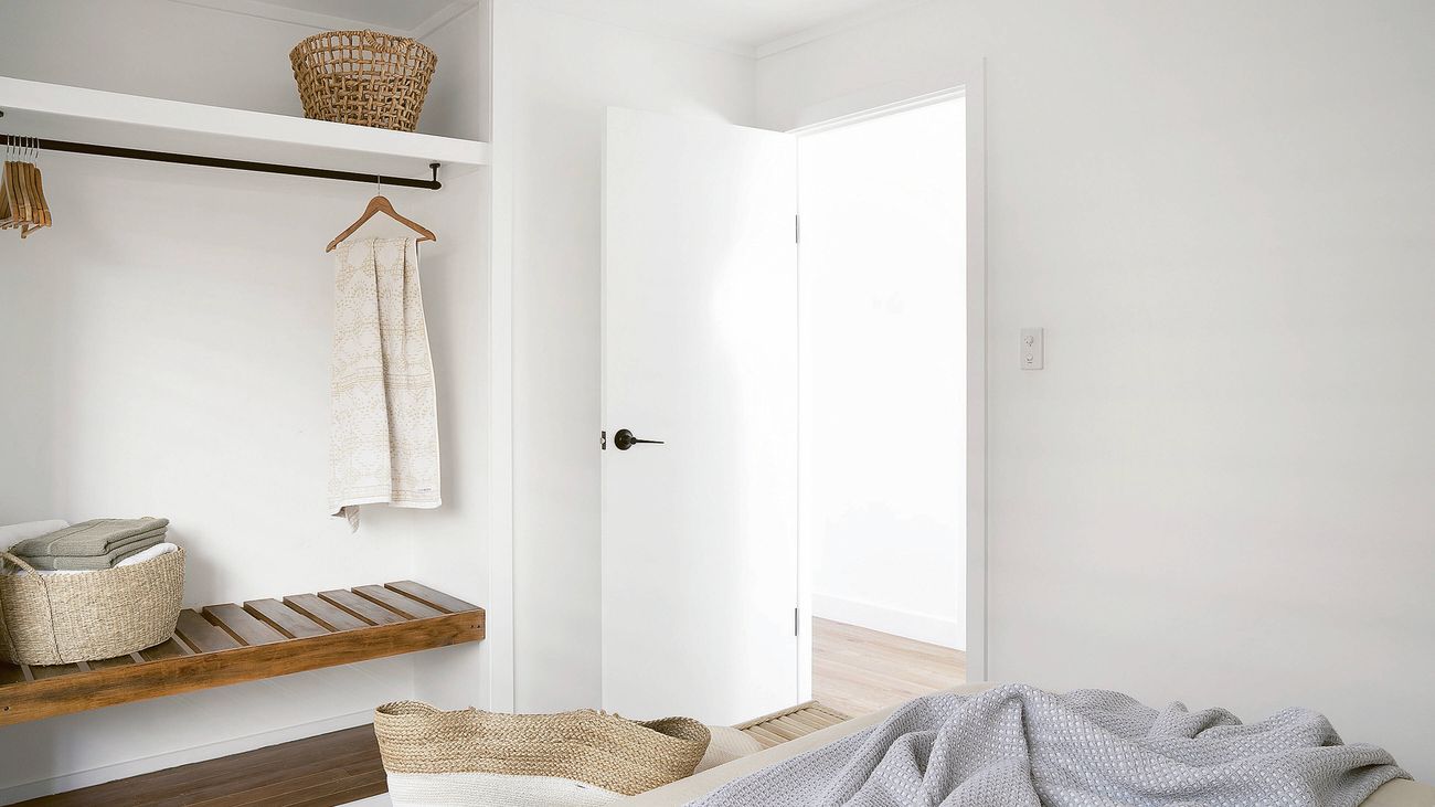 An open-plan white closet with a wooden bench, a hanging rod with a towel, and woven baskets.