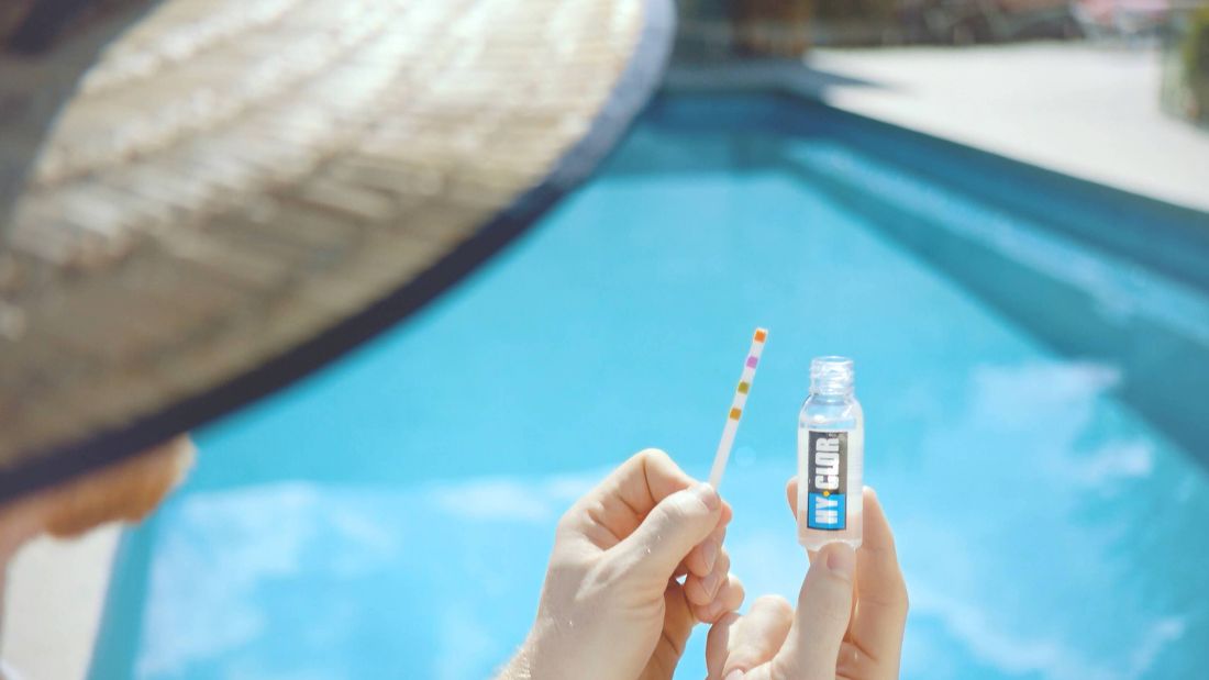 Person holding a swimming pool water testing strip and test solution bottle beside a blue swimming pool.