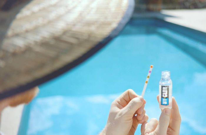 Person holding a swimming pool water testing strip and test solution bottle beside a blue swimming pool.