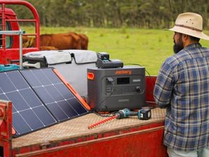 : The back of a male wearing a wide-brimmed hat and a check shirt standing in front of a Jackery portable power station on the back of a ute in a farm setting.