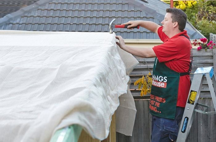 A shade cloth being nailed into place over a wooden frame by a Bunnings team member on a ladder