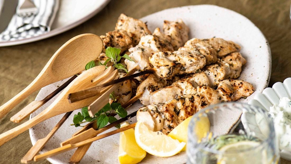 BBQ skewers on a white platter with a wooden serving fork and spoon, lemon wedges and a bowl of tzatziki on the side,