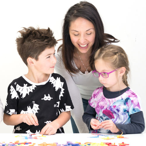 A woman with two young children are at a bench that is covered with mosaic glass tiles. They are smiling and looking at each other. 