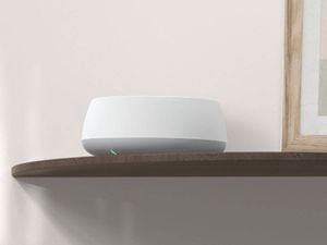 White mesh Wi‑Fi router with a small green status light placed on a wooden shelf against a neutral wall.