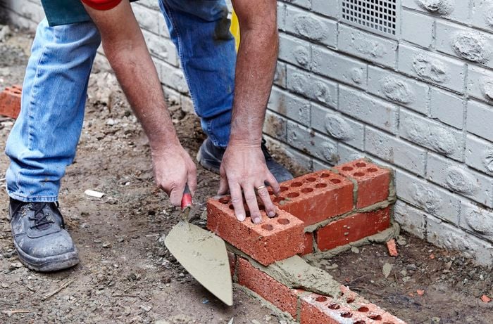 A person laying a second course of bricks parallel to a house wall