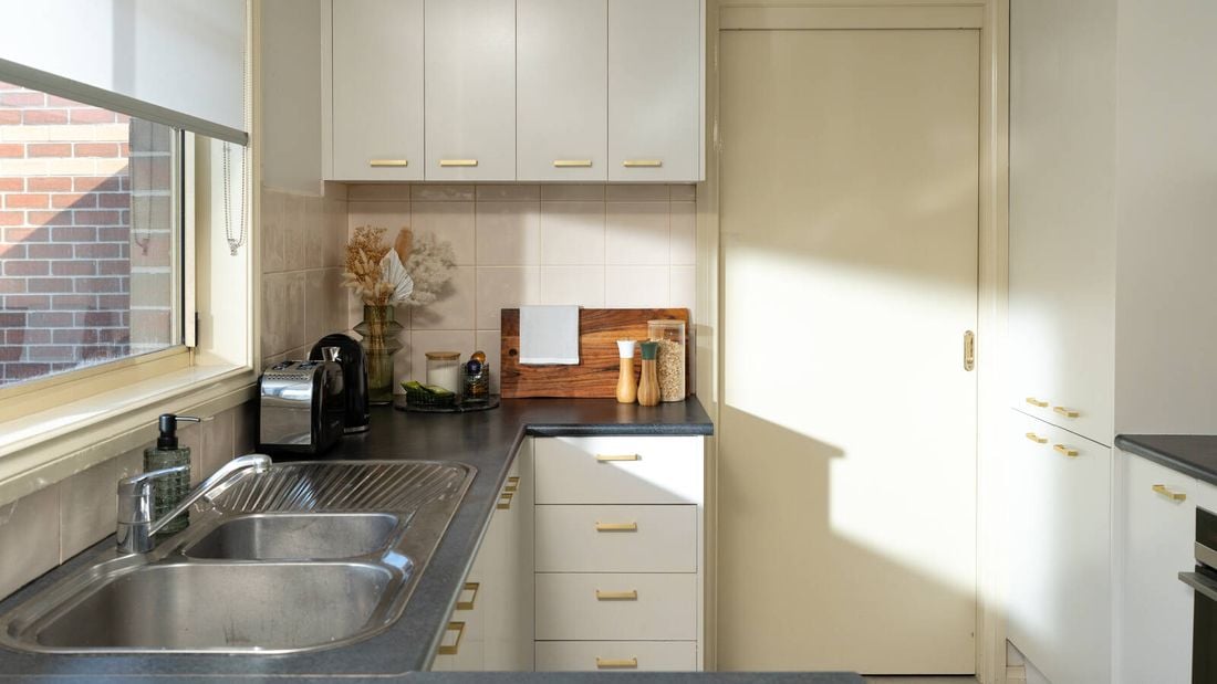 A bright, modern kitchen features white cabinets with gold handles, a double sink, and neatly arranged decor on the countertop