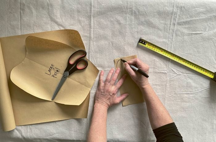 Hands tracing a petal shape onto brown paper with a marker, next to scissors and a tape measure.