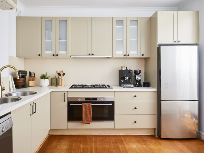 Modern U-shaped kitchen with beige cabinetry, white benchtops, and a built-in oven beneath a gas cooktop, featuring a double sink with a brass tap, small appliances on the counter, and a stainless steel refrigerator.