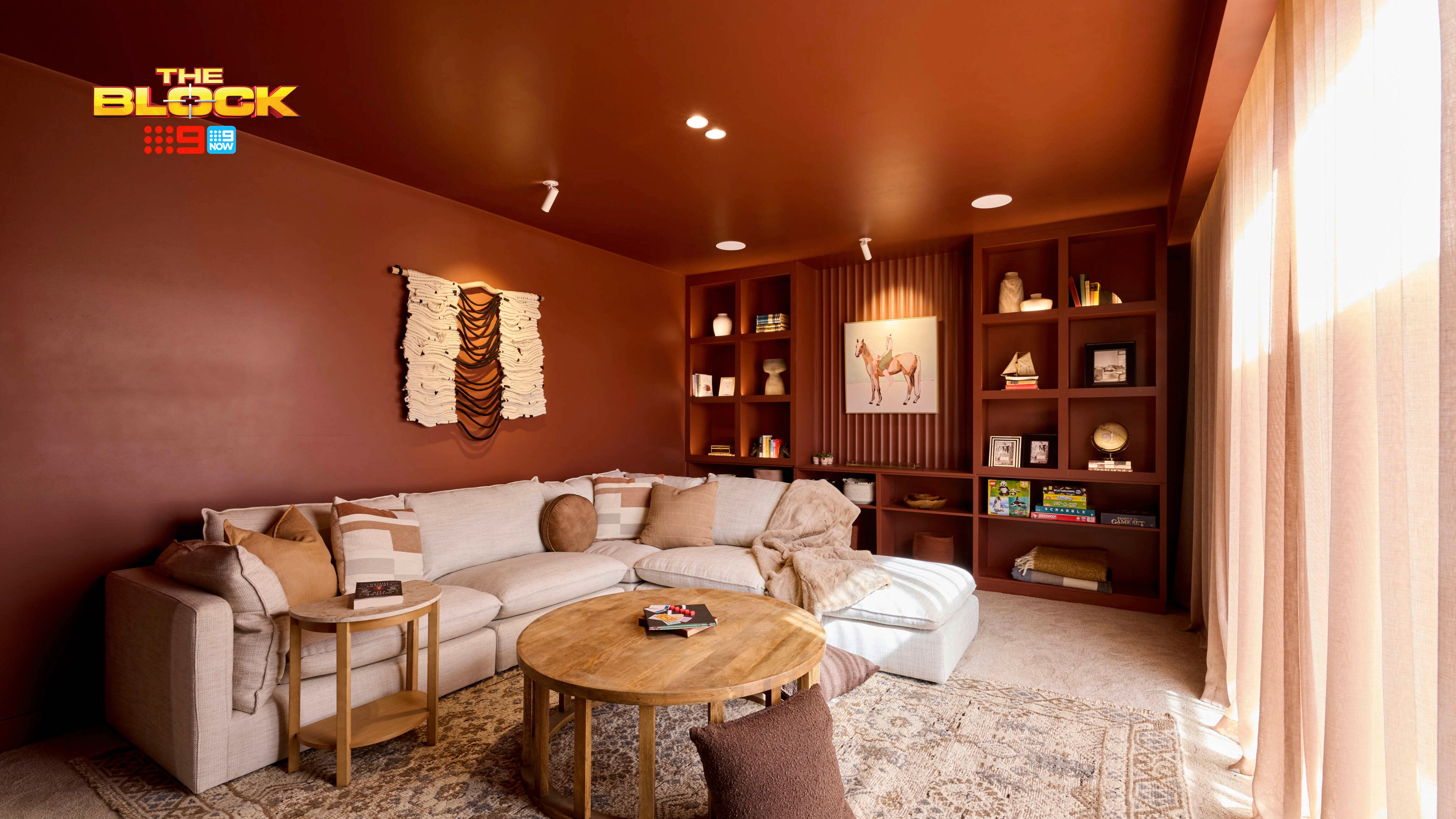 A cozy living room with terracotta-colored walls and ceiling, featuring a large beige sectional sofa adorned with throw pillows and blankets. A round wooden coffee table rests on a patterned rug. To the right, a built-in bookshelf displays books, decorative items, and a horse painting. Natural light filters through sheer curtains on the right side.