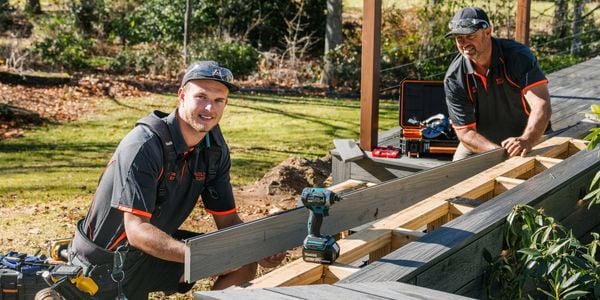 Two tradespeople install grey composite decking on a timber frame in a backyard, one holding a board while the other works nearby. Power tools and a tool case sit on the structure, with grass and trees behind.