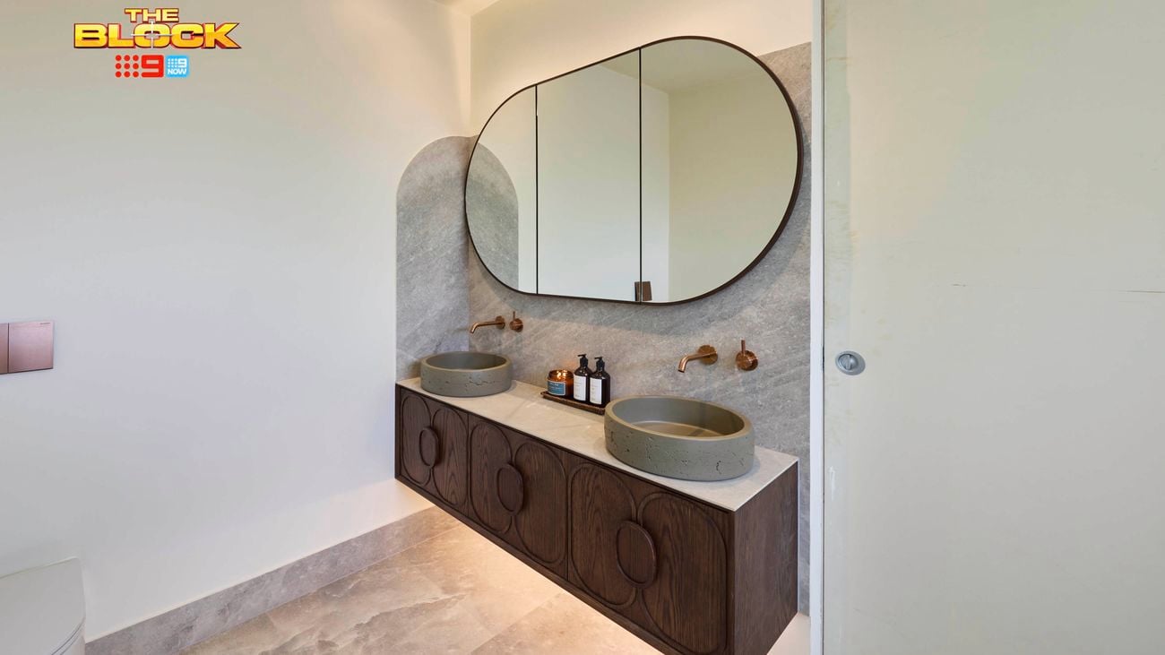Modern bathroom with double stone sinks, brass wall-mounted faucets, oval mirror, marble countertop, dark wood vanity, and light stone tile floor. 