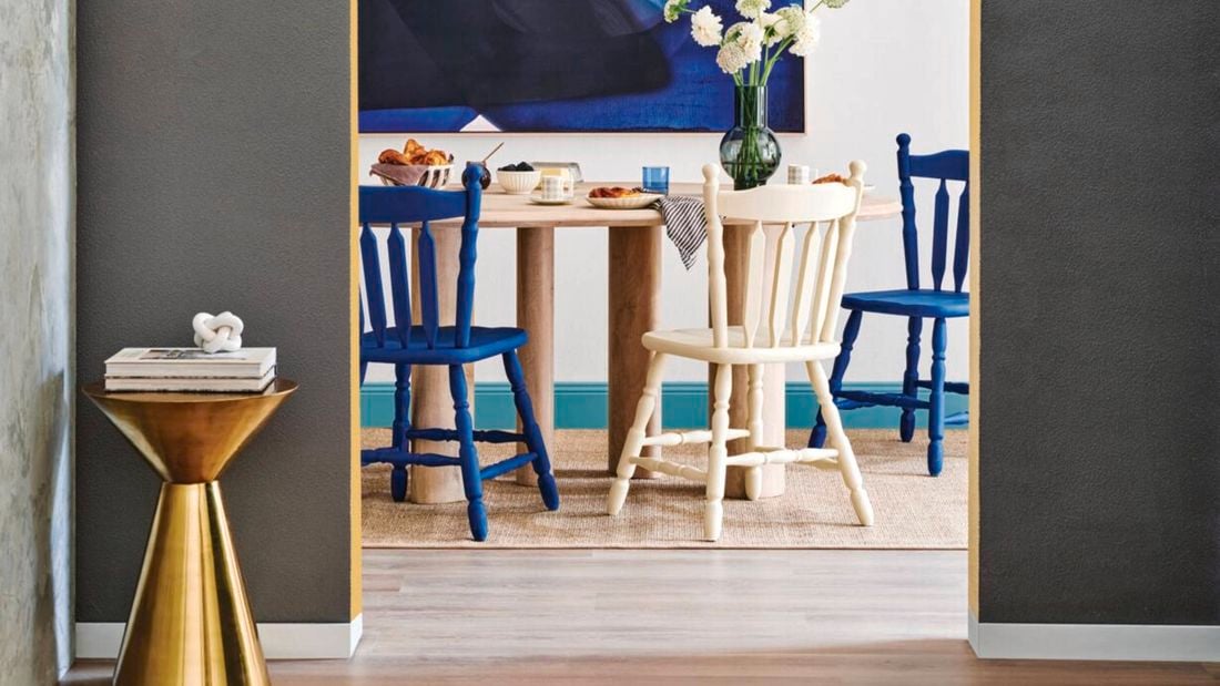 Grey wall with arched doorway, opening out into a white room with a blue abstract painting and a wooden dining table with blue and white chairs.