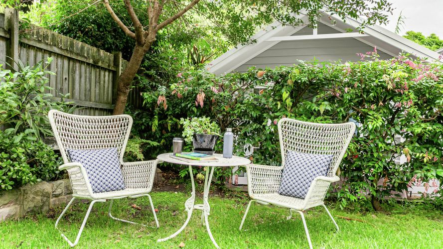 Two white woven outdoor chairs with blue cushions and a small round table set on a grassy garden lawn, surrounded by leafy trees, flowering shrubs and a timber fence.