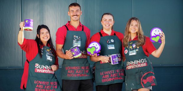 Four Bunnings team members wearing red shirts and aprons smiling while holding colourful circular awards and a paint tin against a dark blue background.