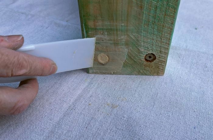 A hand using a white scraper to smooth wood filler on a piece of timber