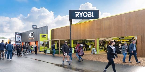 People walking along a wet pathway at an outdoor trade show, passing a large RYOBI display booth with bright green branding and tool displays, alongside a neighbouring AEG Powertools stand, under a partly cloudy sky.