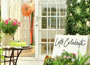 An outdoor patio with a bistro table, a planter of flowers, and a white door with a large paper flower decoration.