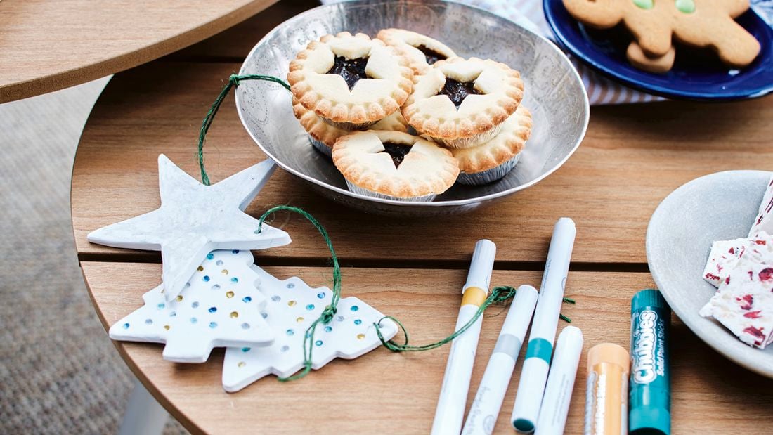 esting round wooden tables with craft supplies: white star and tree ornaments with green string, textas and a glue stick. A metal bowl of jam tarts and a blue plate with gingerbread biscuits sit on the tables, with a small bowl of nuts behind.