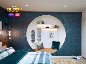 Blue bedroom with a double bed, textured blue wallpaper and blue checked linen. The bed is on a brown checked rug and there is a circular recess in the wall with a bench seat and clothing hooks.