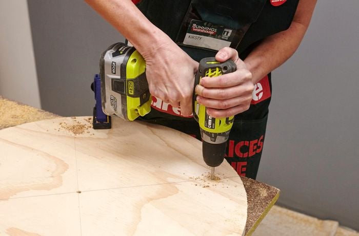 A person drilling a hole in a round sheet of plywood using a cordless drill