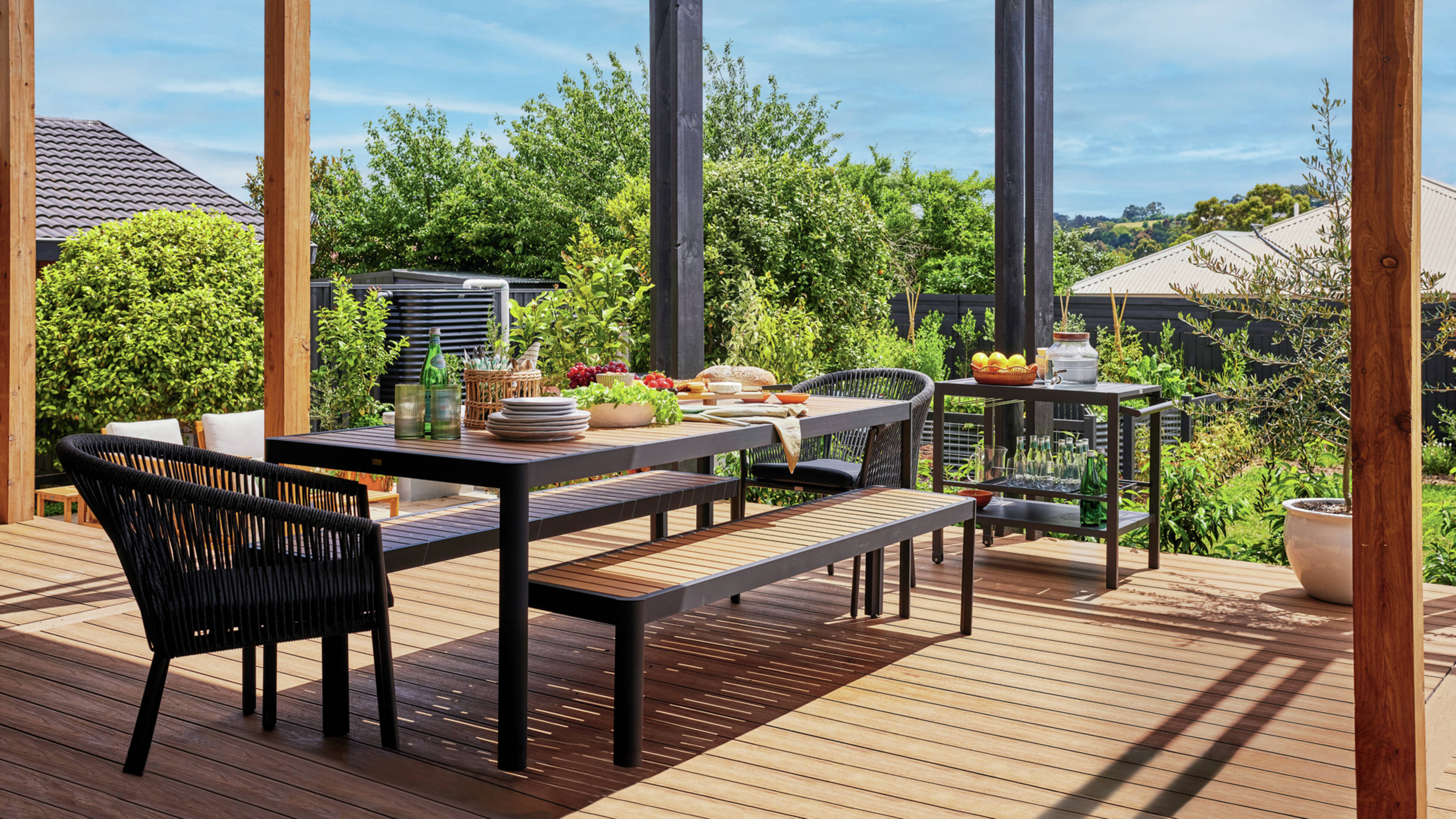 Outdoor deck with dining table set for a meal under a pergola, surrounded by greenery.