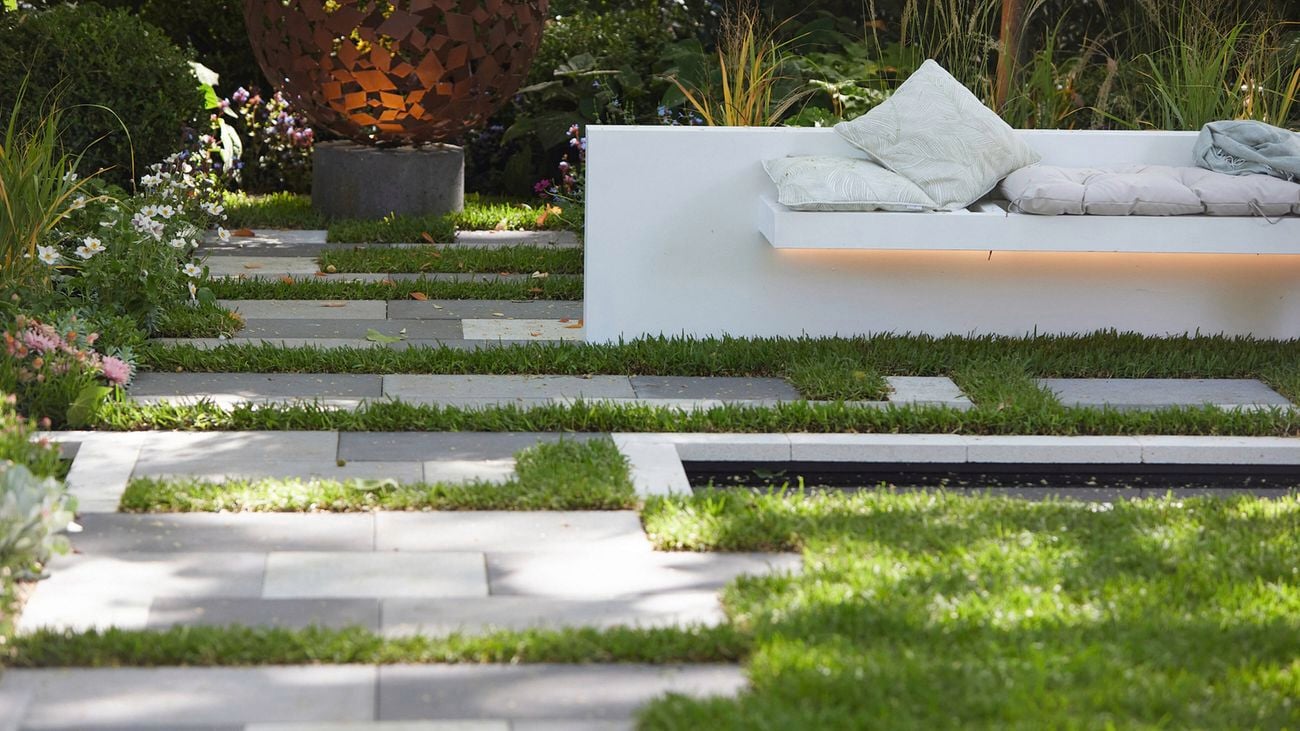 A garden with two-tone stone paving with grass, a still rectangular pond and a built-in stone bench with large cushions on top. In the background is an oversized metal globe sculpture on a stone plinth.