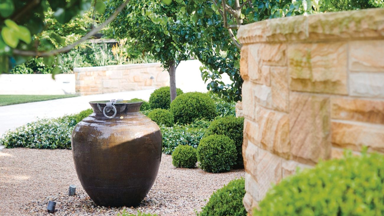 Garden with a large pot water feature, surrounded by trimmed bushes, trees, and a cobblestone ground.