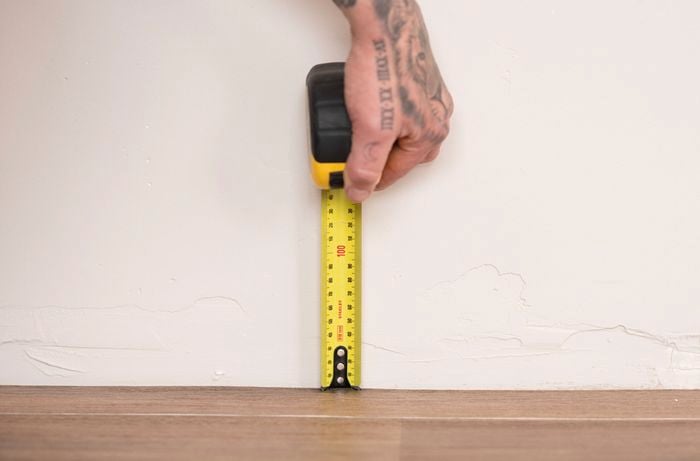 A person is using a measuring tape to measure the gap between the skirting board and the floor to ensure it fits the carpet's thickness.