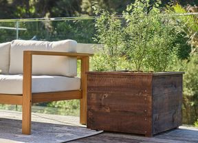 A wooden planter box with green plants next to an outdoor sofa on a deck.