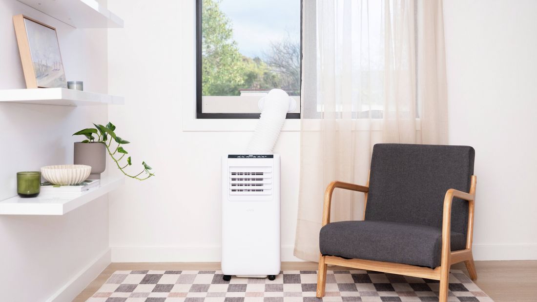 Portable air conditioner positioned beneath a window in a bright living room, with an exhaust hose vented outside. A dark grey upholstered armchair sits beside the unit, while floating shelves with a potted plant and decorative items line the wall. A patterned rug covers the floor and sheer curtains frame the window.