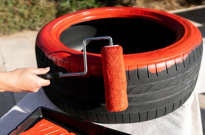 A person is applying red paint to an old tyre using a paint roller outside.