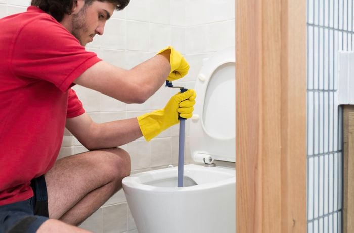 A person wearing a red shirt and yellow rubber gloves is using a drain auger to unclog a toilet in a tiled bathroom.