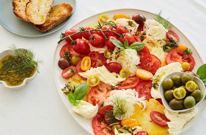 Red and green tomatoes, fennel and other various vegetables make a tomato carpaccio salad with slices of bread next to it.