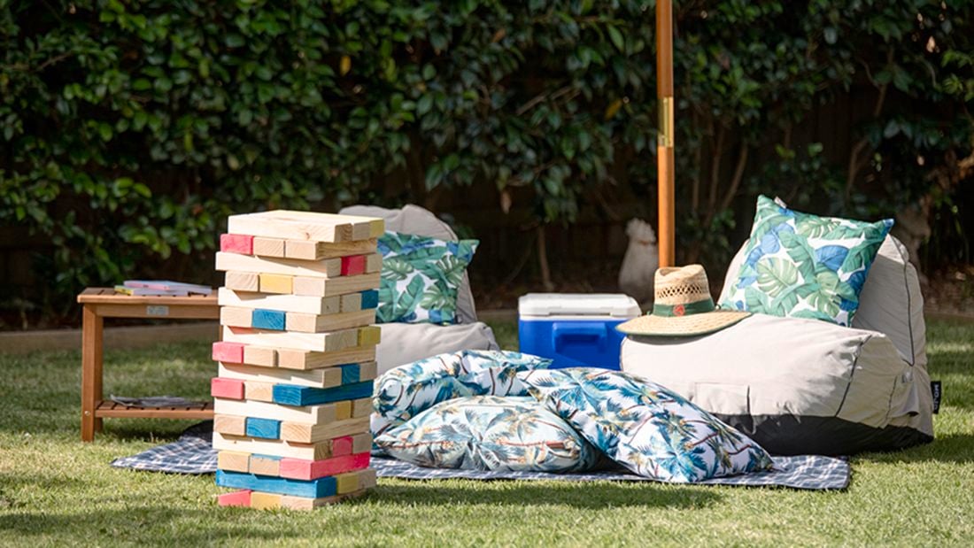 Giant completed tumbling tower game stacked up in an outdoor setting with large cushions, table, hat and esky under an umbrella