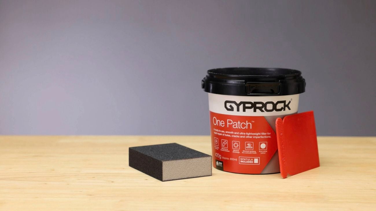 A tub of Gyprock One Patch on a table with a spatula and a sanding block, in front of a grey wall