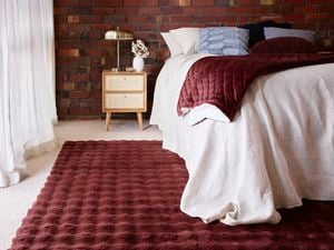 A bedroom with a brick wall, bed, side table with a lamp on it and a burgundy rug on the floor in it.
